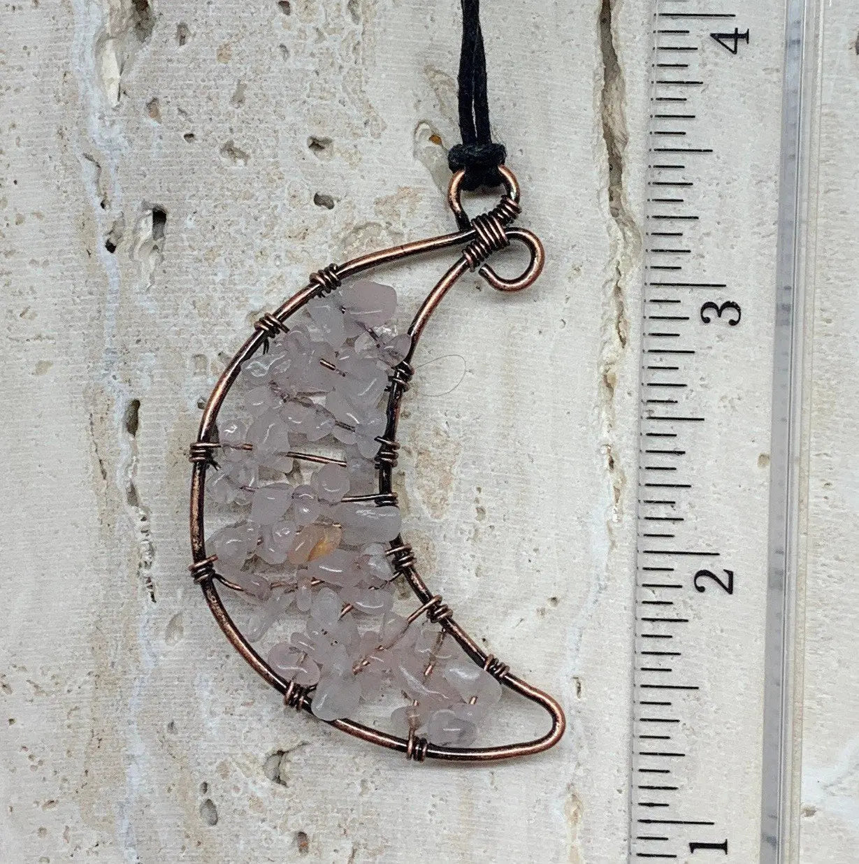 Rose quartz chip beads intricately woven into a copper wire wrapped crescent moon amd attatched to an adjistable black cord, displayed next to a ruler. pendant 2" long.