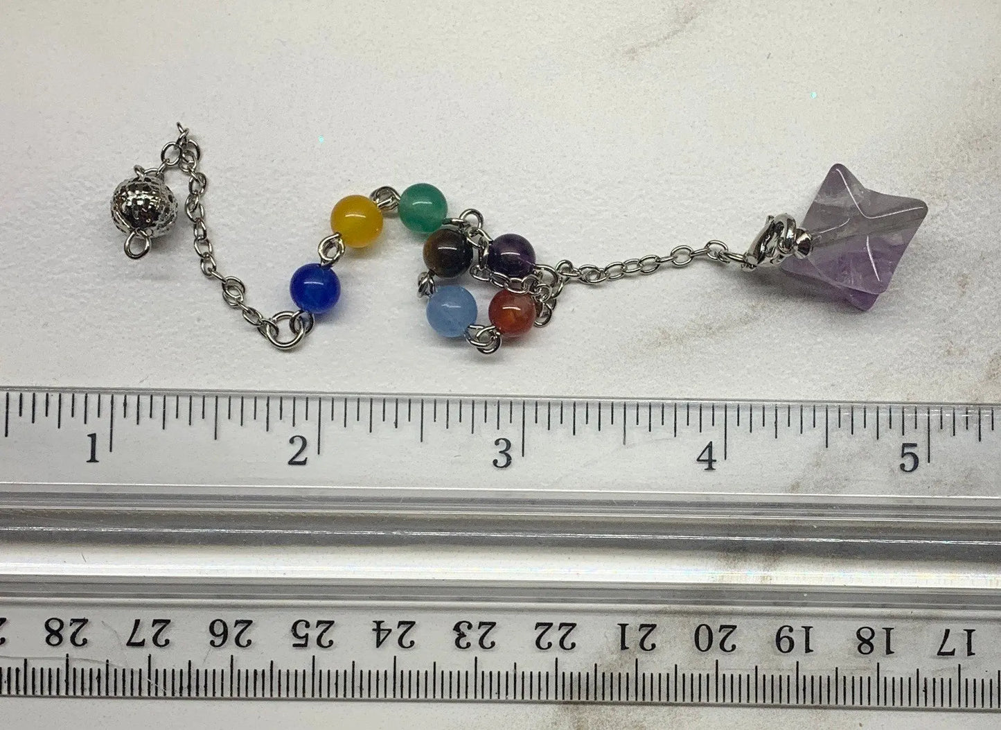 3/4 inch amethyst merkaba pendulum attatched to an 8 inch silver chain containing chakra beads displayed next to a ruler