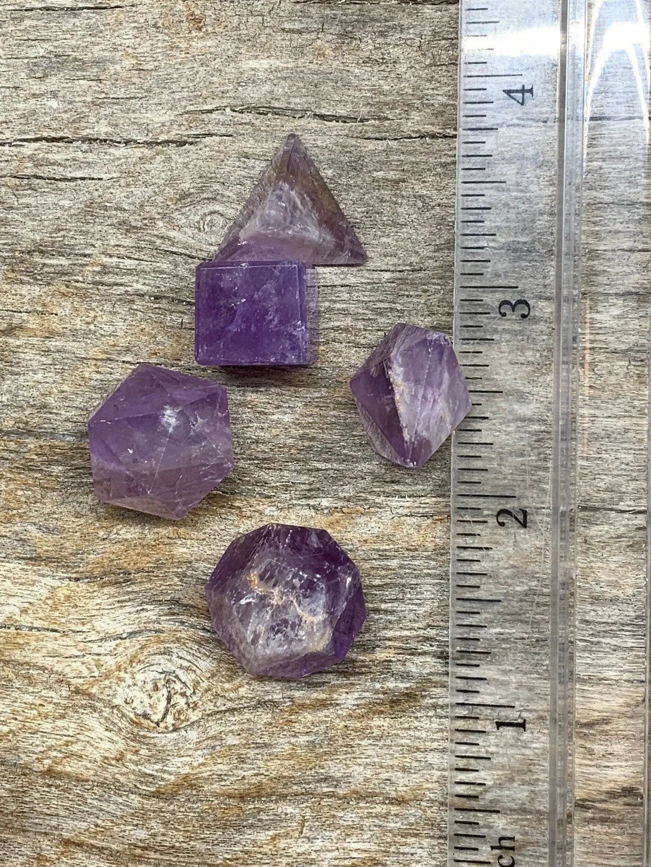 Collection of 5 amethyst crystal shapes including pyramid, cube, octahedron, decahedron, and dodecahedron, displayed next to a ruler. Each crystal is approximately 1/2” in diameter.