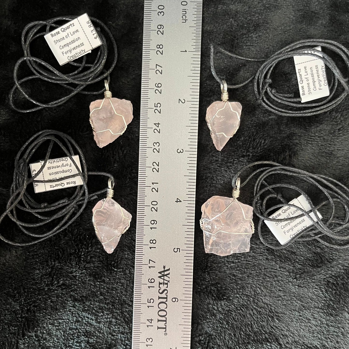 4 Handcrafted wire-wrapped rose quartz necklaces featuring polished crystal of various sizes ntricately woven with silver wire and artatched to adjustable black cords, displayed mext to a ruler. Rose quartz crystals are 3/4" -1" long