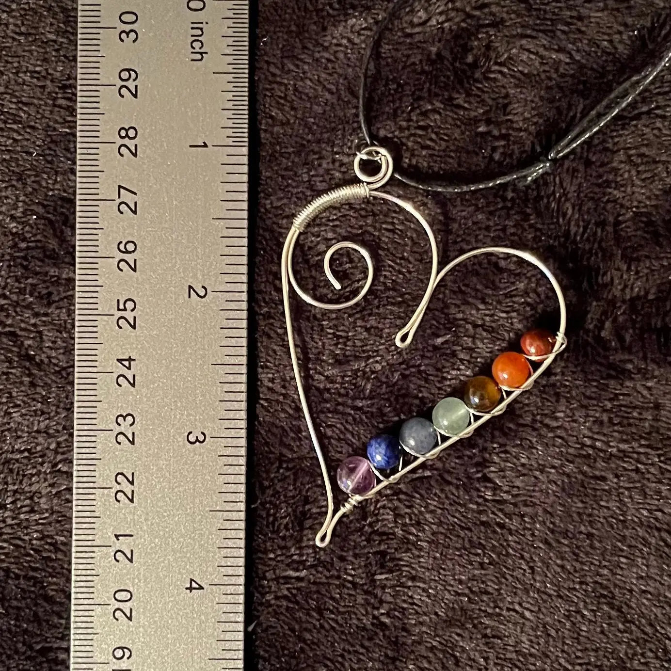 7 chakra beads spiral wire wrapped with silver wire in a line along the inside edge of a silver heart. attatched to a an adjustable black cord, displayed next to a ruler to show size. pendant is approximately 2 1/2 inches long