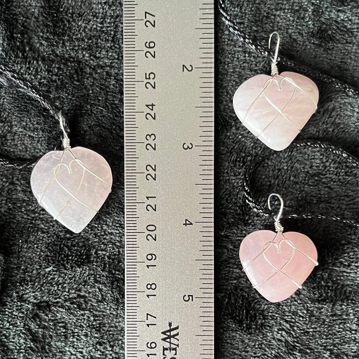 Three Wire-wrapped rose quartz heart necklaces: a silver wire intricately wraps around polished rose quartz heart pendants, hanging on a delicate chain, displayed next to a ruler. hearts arw approximately 1" long