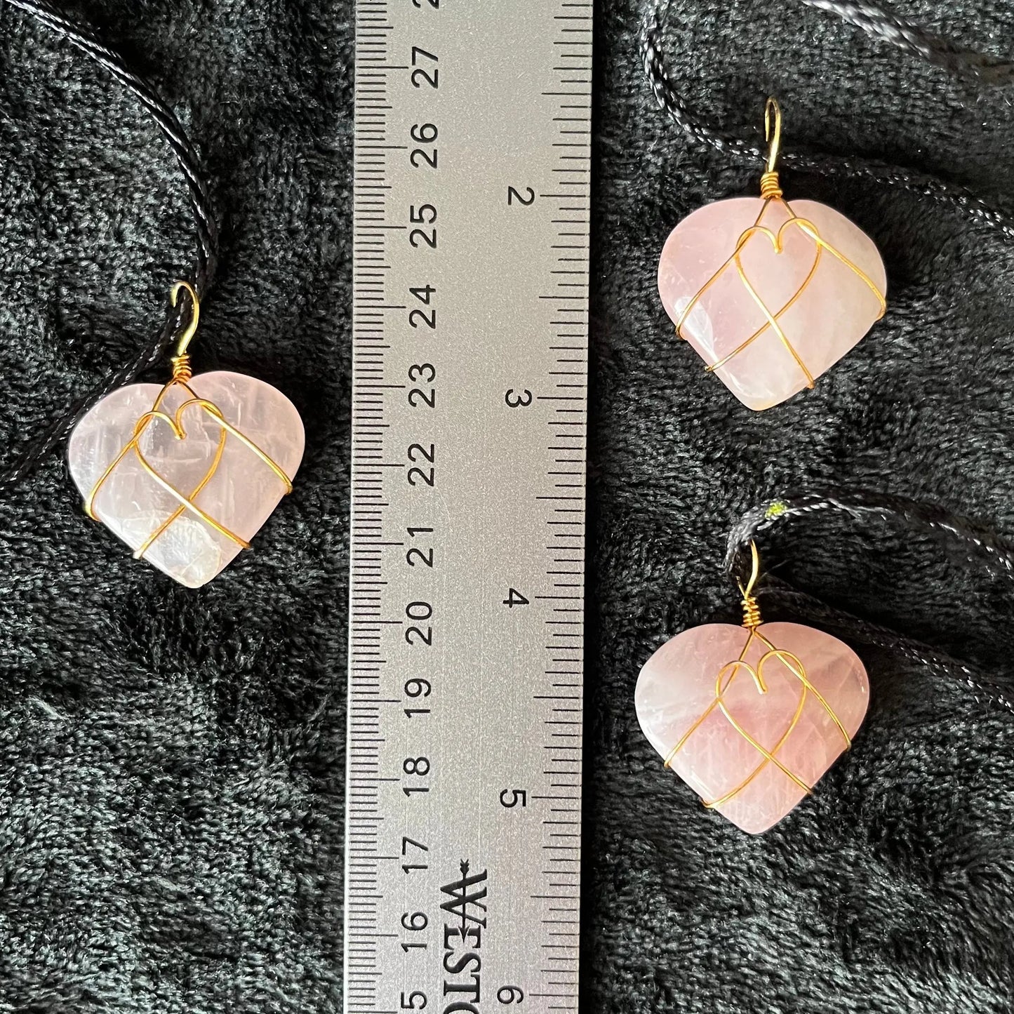 Three wire-wrapped rose quartz heart necklaces: a gold wire intricately wraps around polished rose quartz heart pendants, hanging on a delicate chain, displayed next to a ruler. hearts are approximately 1" long