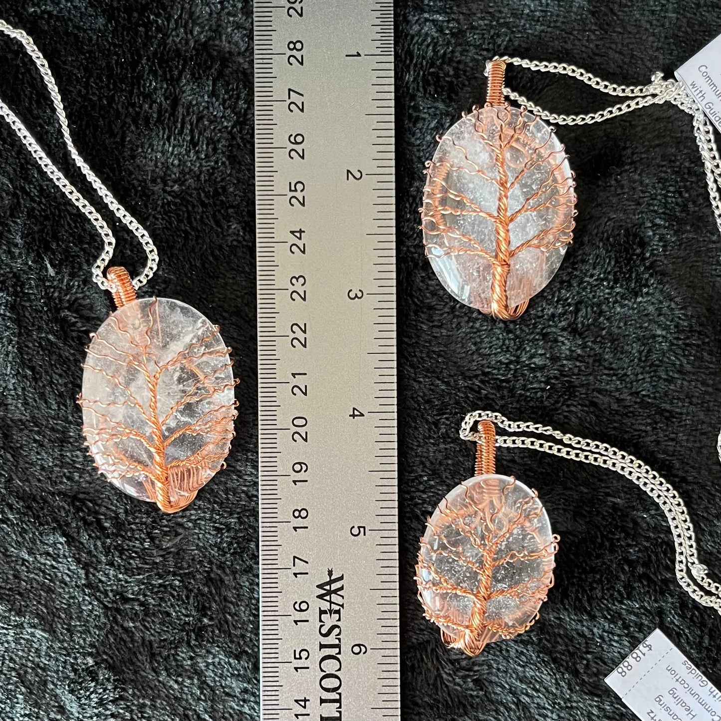 Three tree of life copper wire wrapped clear quartz oval stone pendants attatched to a silver chains, displayed against a black background next to a ruler. eaxh oendant is 1 3/4" - 2 1/4" long