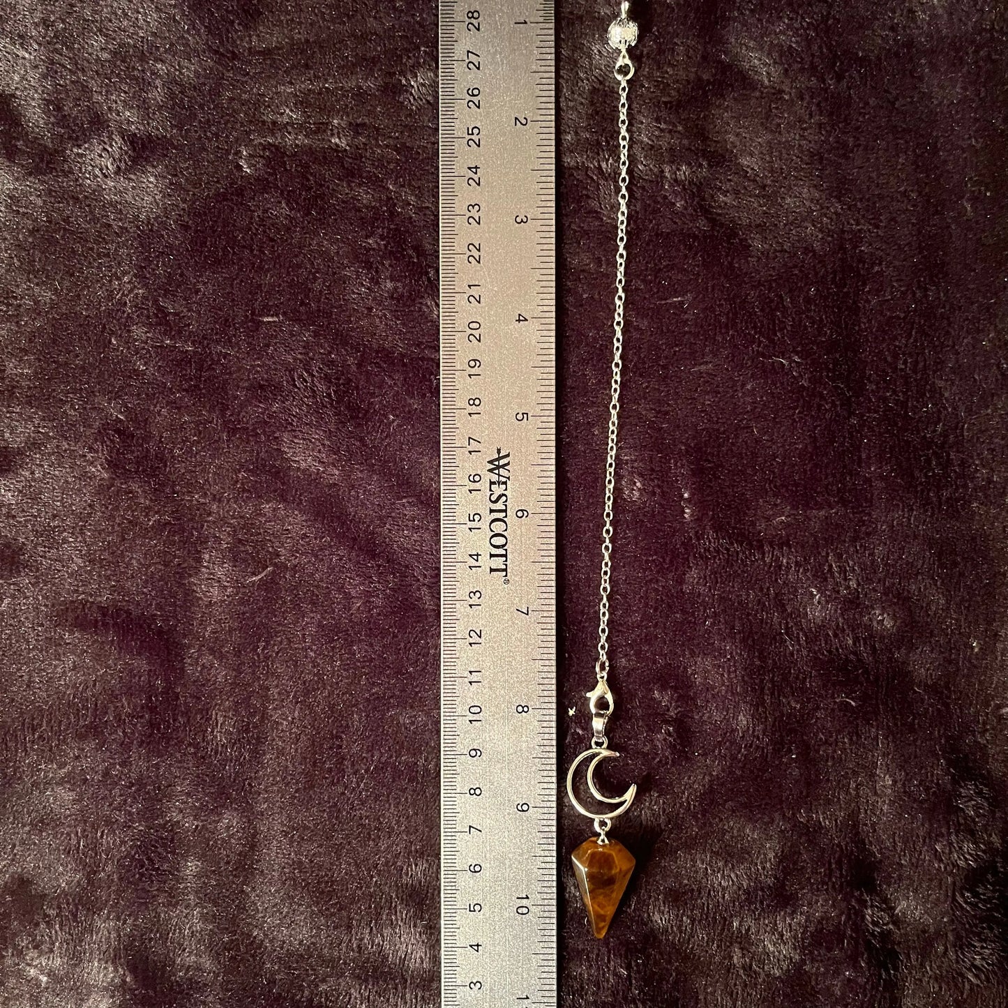 close up of a 1 inch bronze and brown marbled tiger eye pendulum suspended from a silver creacent moon charm attatched to a silver 8 inch chain displayed next to a ruler
