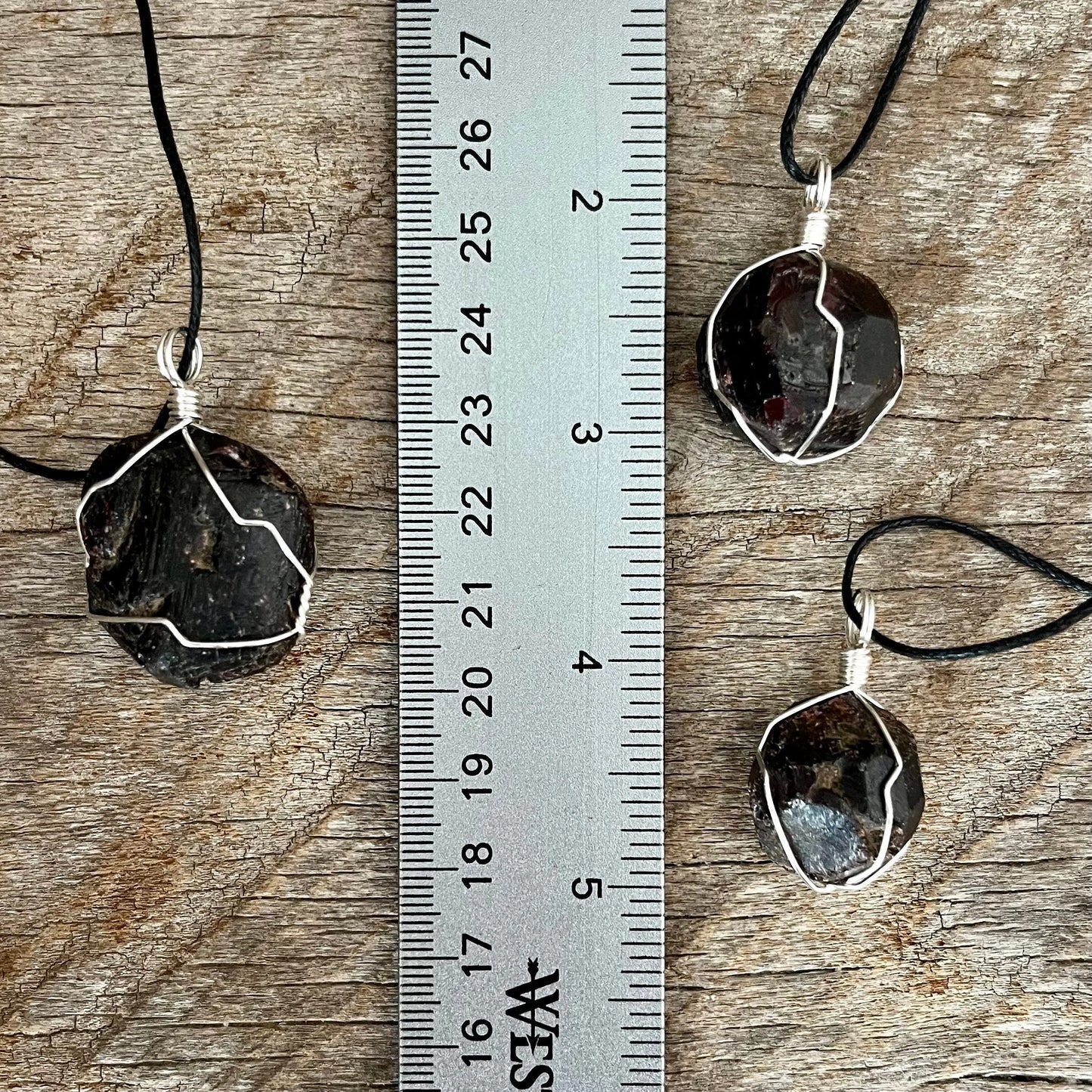 3 silver wire wrapped garnet crystal pendants attatched to adjistable black cords, displayed next ro a ruler. pendant vary in diameter from 1/2" to 3/4",