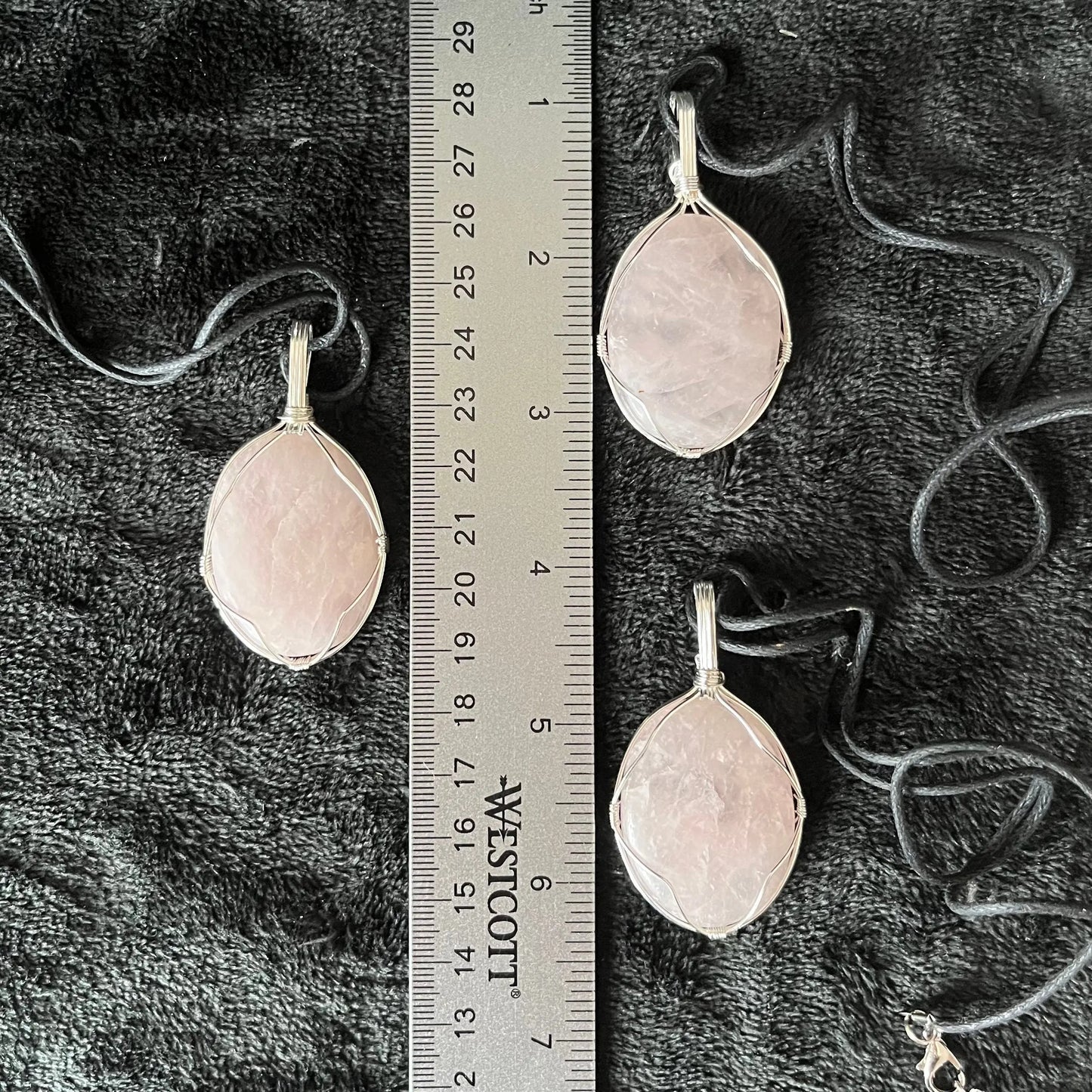 Silver wire wrapped with ornate and delicate frames, these translucent pink rose quartz worry stone pendants are approximately 1 1/2" long, and attached to adjustable black cords and displayed next to a ruler.