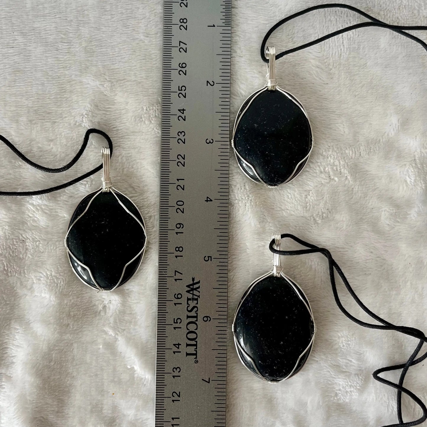 3 obsidian worry stones beautifully framed by silver wire and made into pendants, with adjustable black cords, displayed next to a ruler. obsidian crystal worry stones are approximately 1 1/2" long