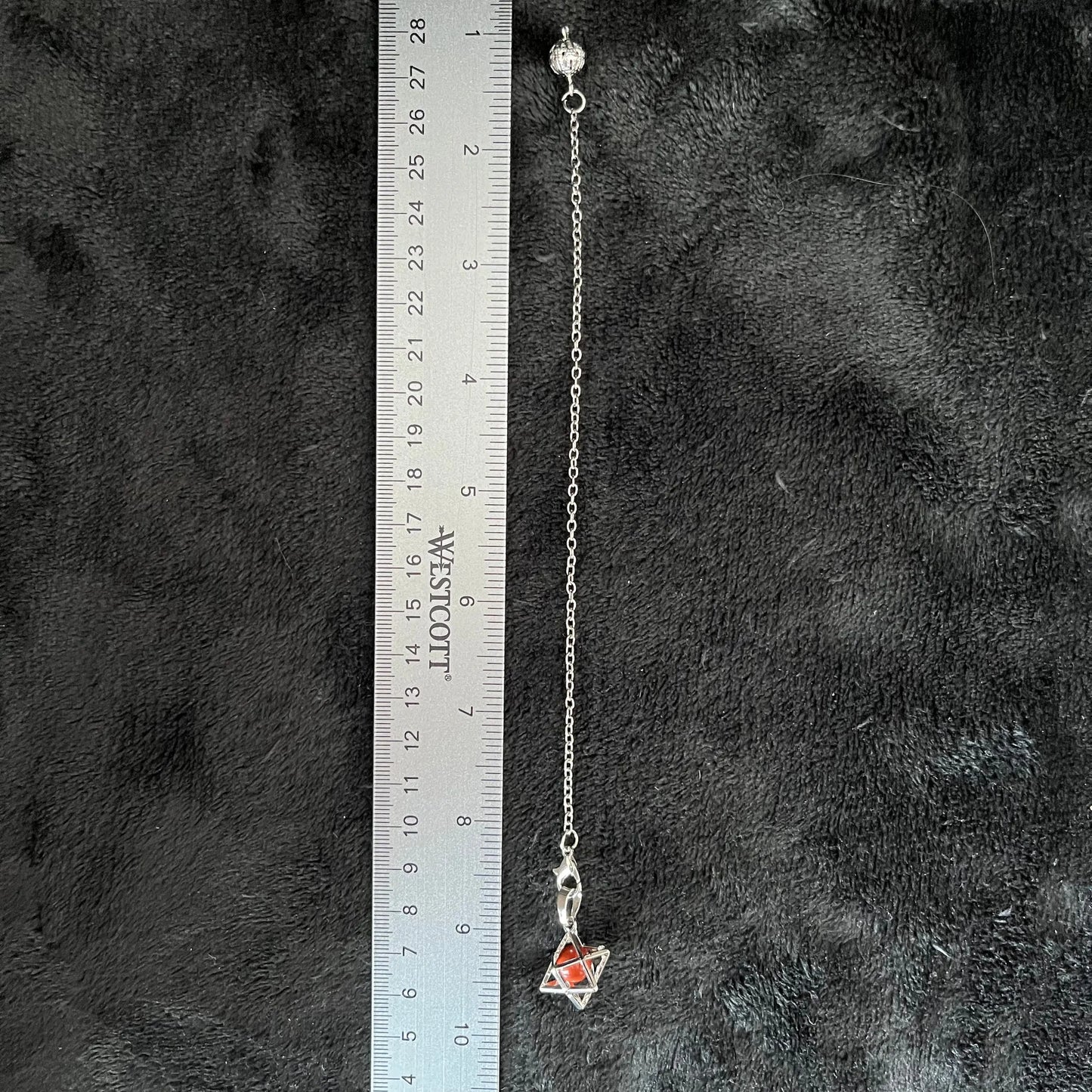 "Close-up of a red Jasper sphere encased within a silver Merkaba pendulum, next to a ruler