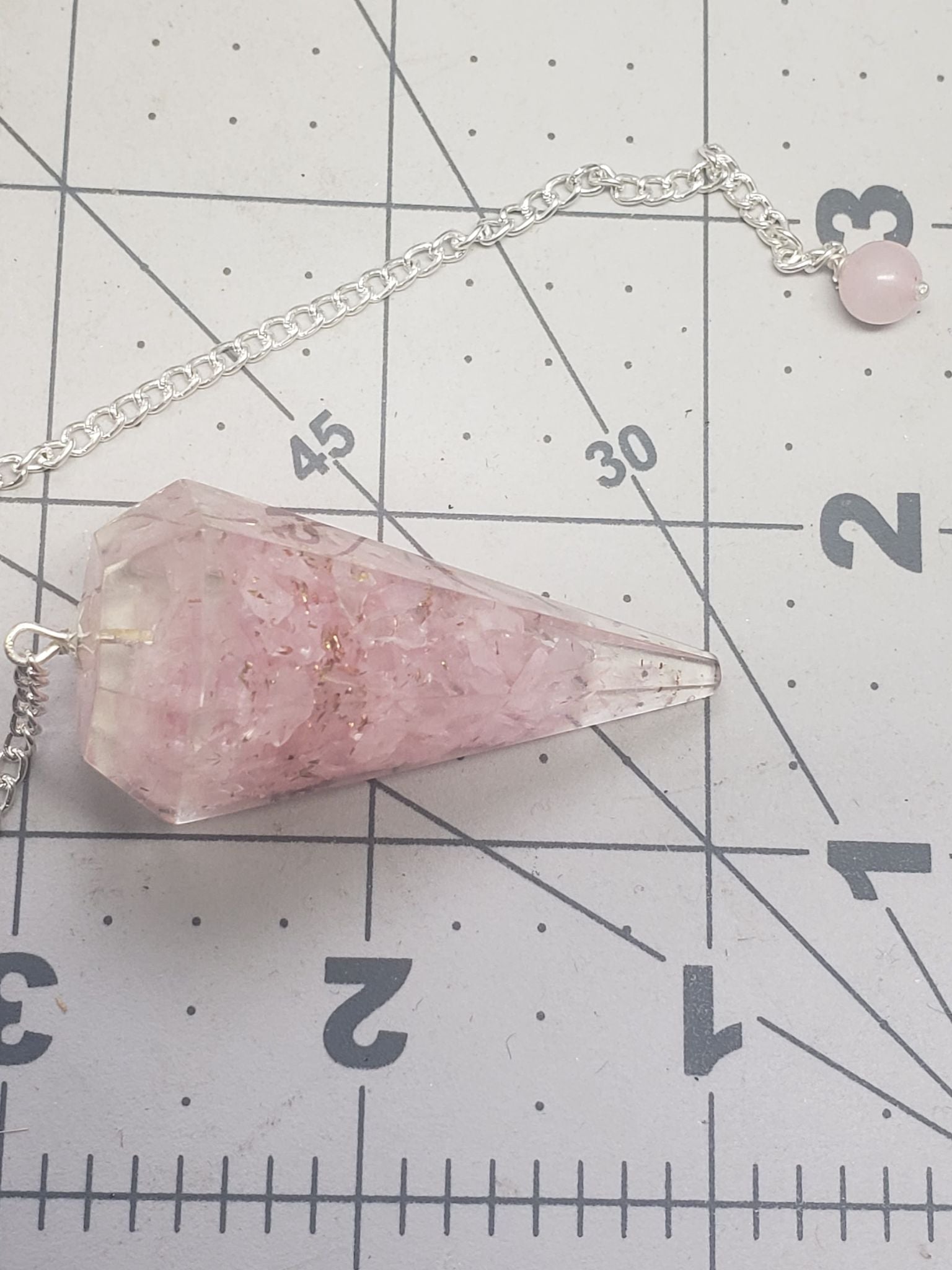 close up of 2 inch rose quartz orgonite pendulum including an 8 inch silver chain, displayed next to a ruler to show size