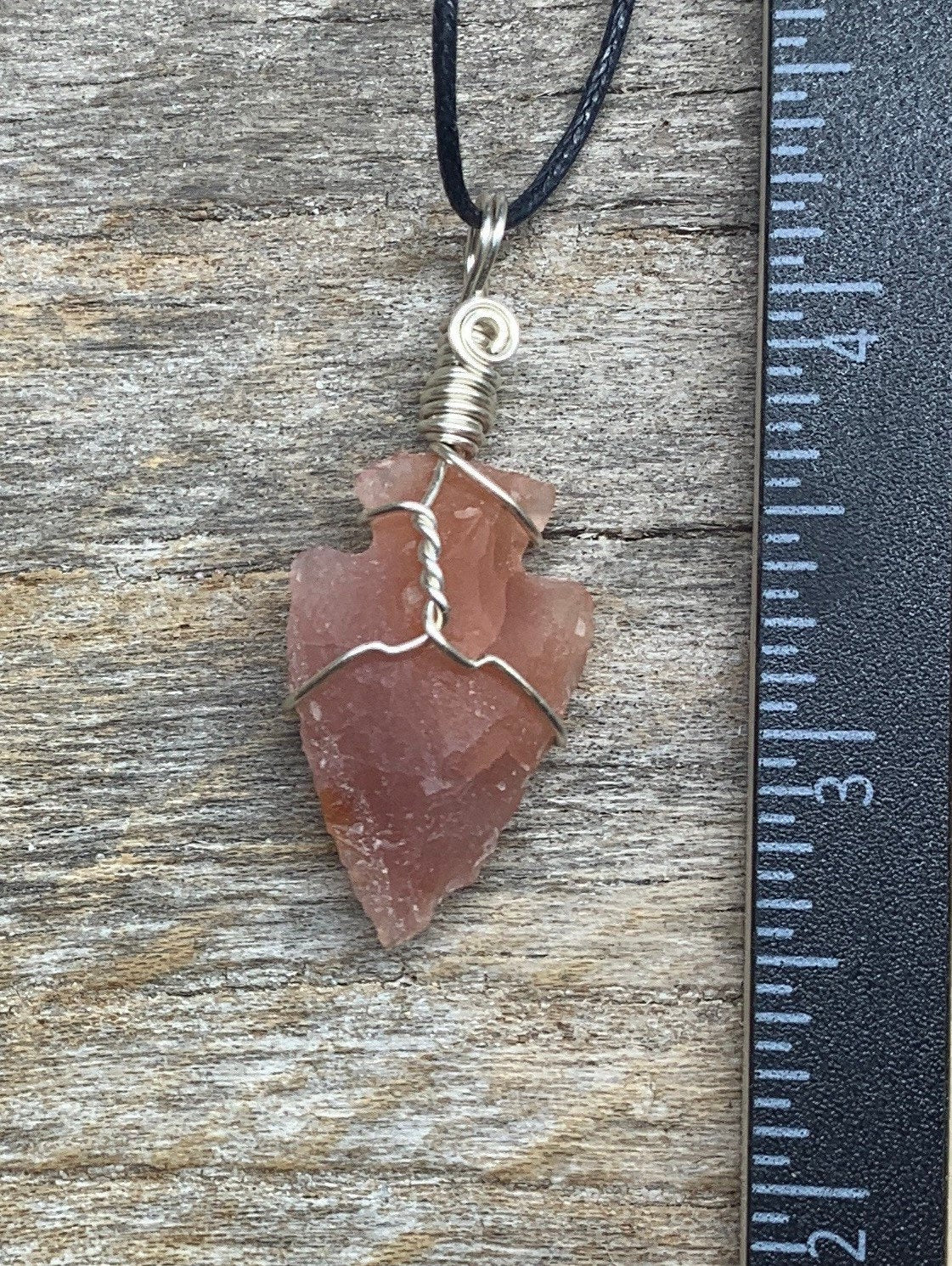 1 inch vibrant orange raw carnelian agate arrowhead, handmade, silver wire wrapped necklace, attatched to an adjistable black cord. lying next ro a ruler