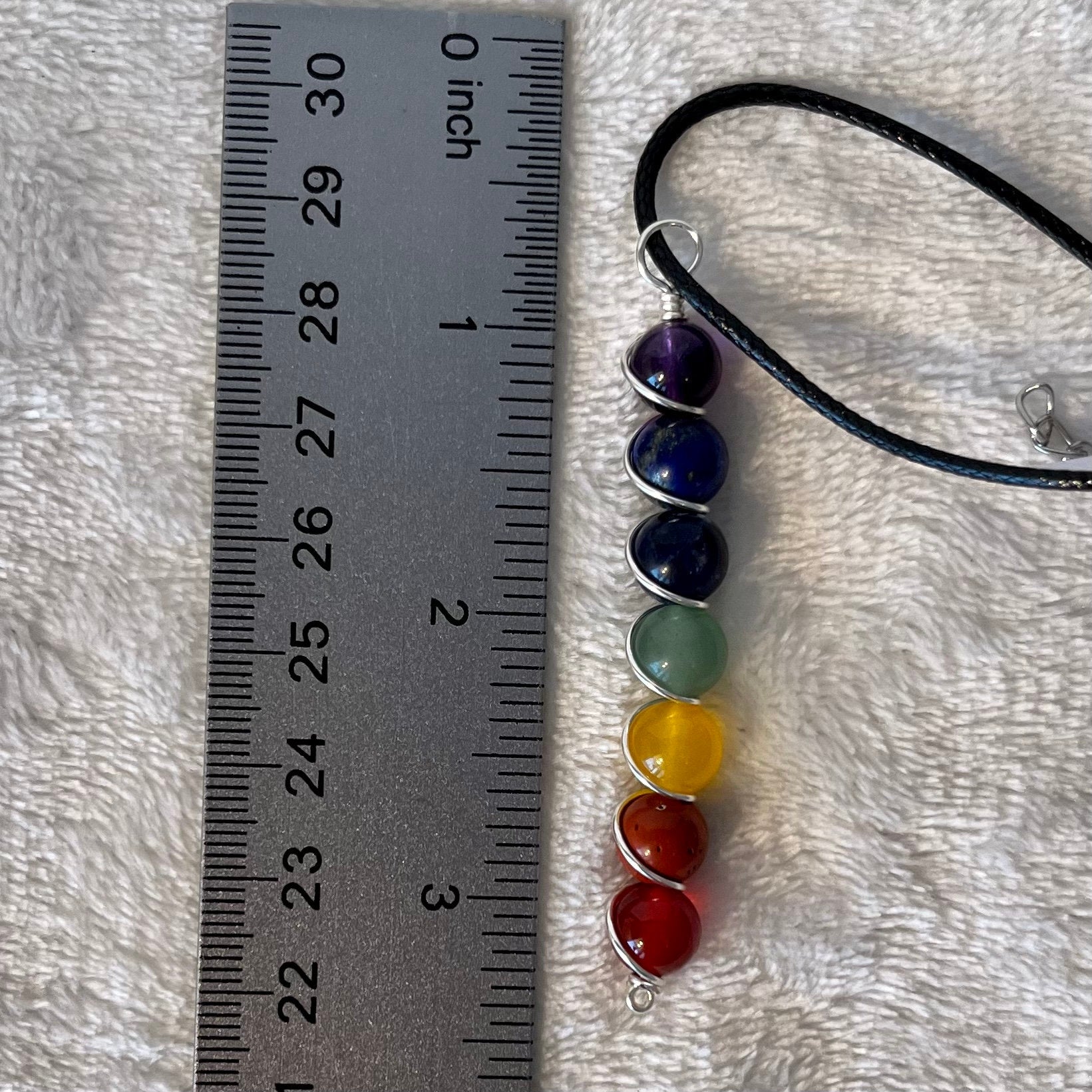 7 chakra beads spiral wire wrapped with silver wire in a line, attatched to a black cord displayed next to a ruler showing pendant is 2 1/4 inch long.