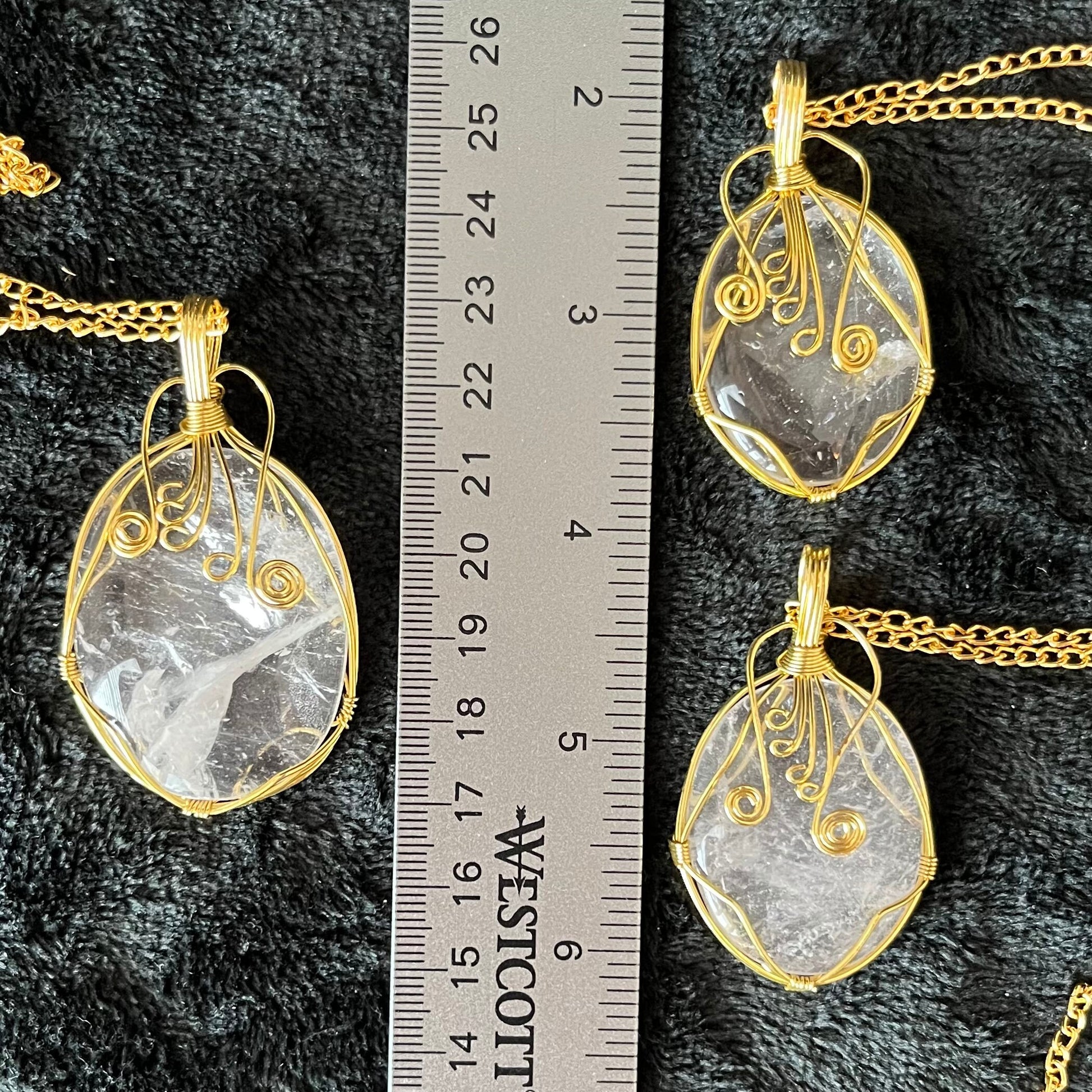 Three gold fancy wire wrapped clear quartz oval stone pendants attatched to gold tone chains, displayed next to a ruler.  stone range ffrom 1 1/4" - 1 1/2" long.