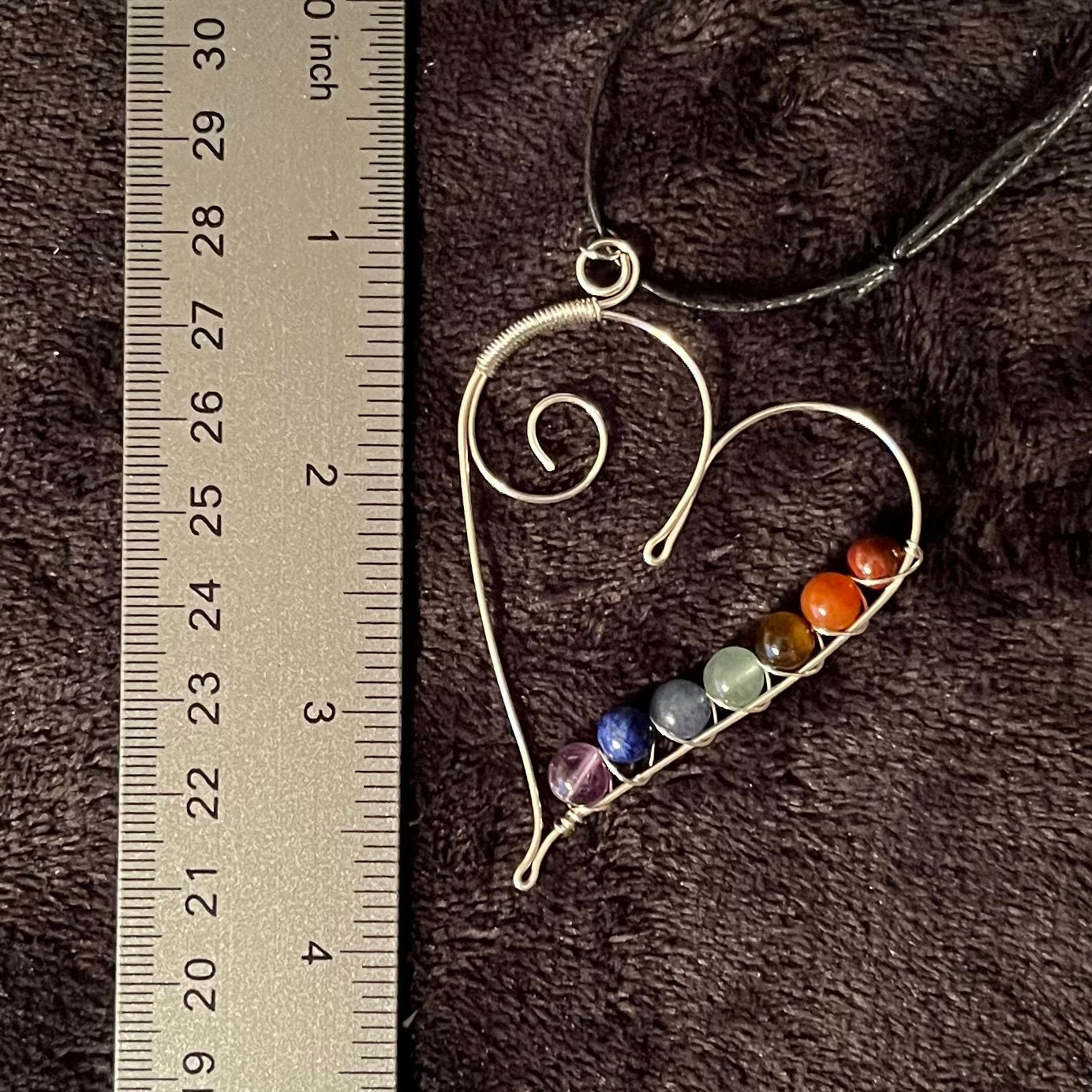 7 chakra beads spiral wire wrapped with silver wire in a line along the inside edge of a silver heart. attatched to a an adjustable black cord, displayed next to a ruler to show size.   pendant is approximately 2 1/2 inches long