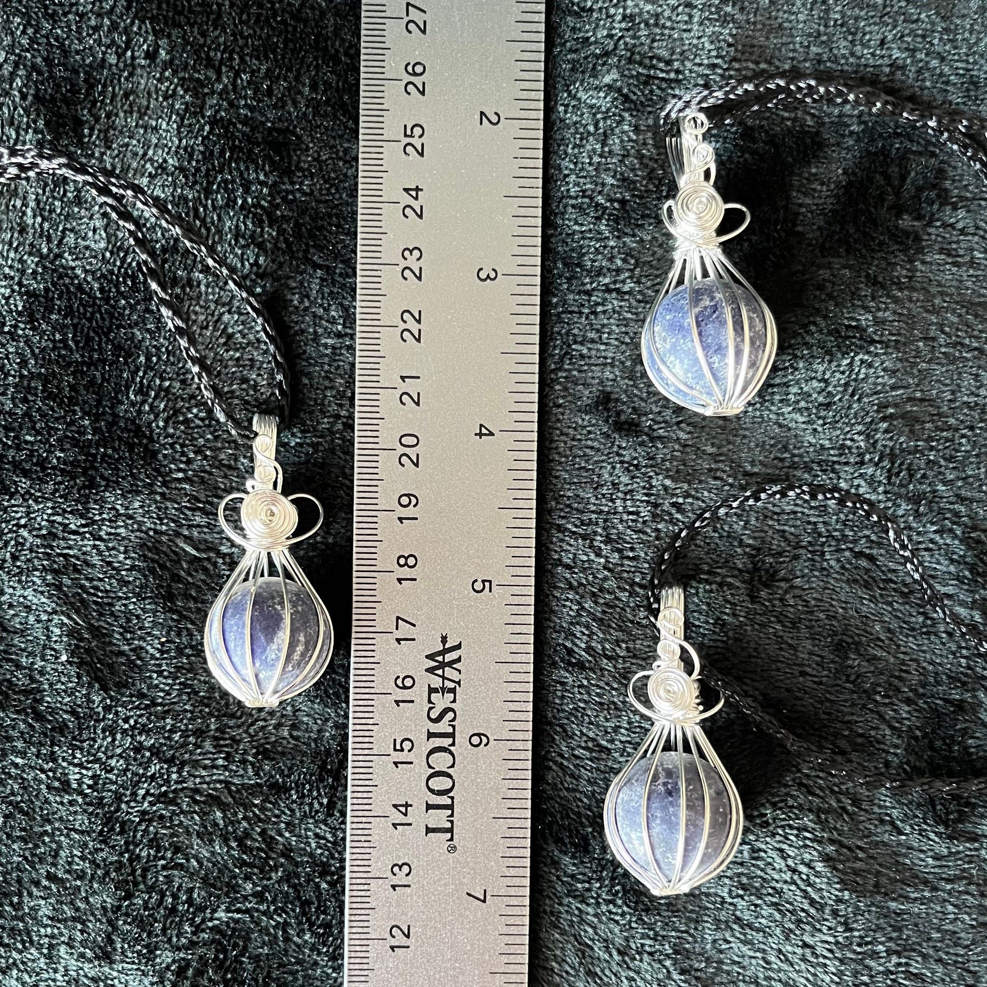 Ornately silver wire wrapped basket pendants containing a blue amd grey sodalite spheres approximately 1/2" - 3/4" in diameter and attatched to black cords, displayed next to a ruler