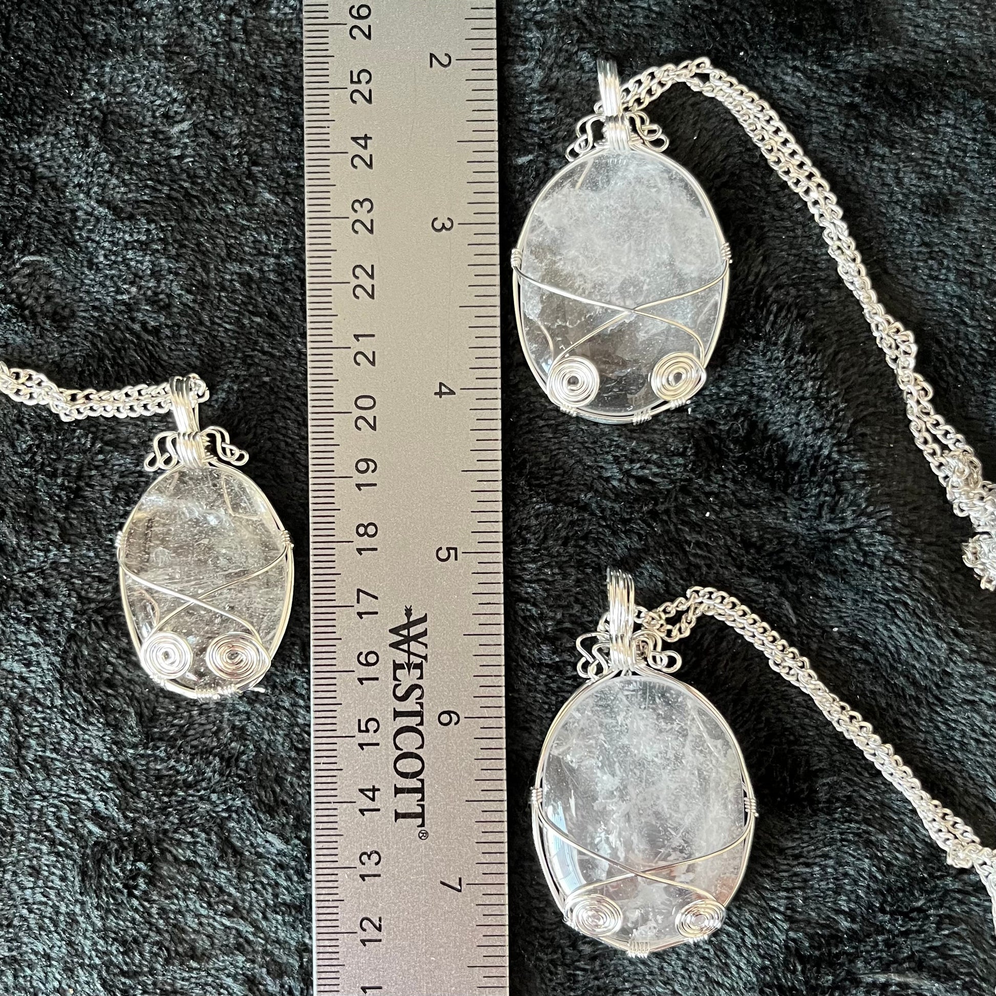 Three silver fancy wire wrapped clear quartz oval stone pendants attatched to a silver chains, displayed against a black background next to a ruler.  eaxh oendant is 1 3/4" -  2 1/4" long