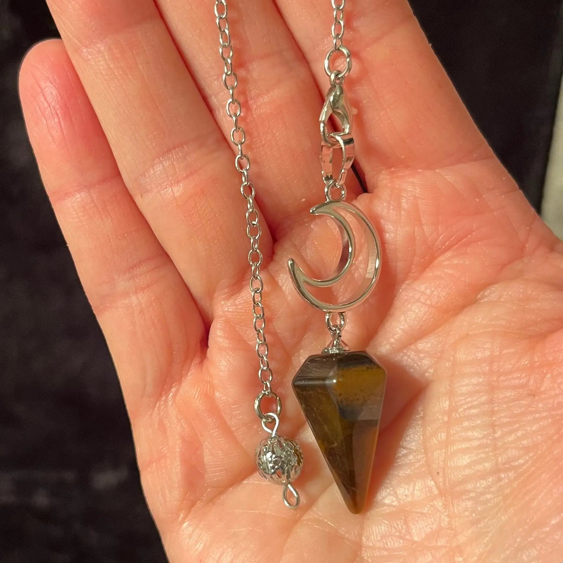 close up of a 1 inch bronze and brown marbled tiger eye  pendulum suspended from a silver creacent moon charm attatched to a silver 8 inch chain held delicately in the palm of a hand.