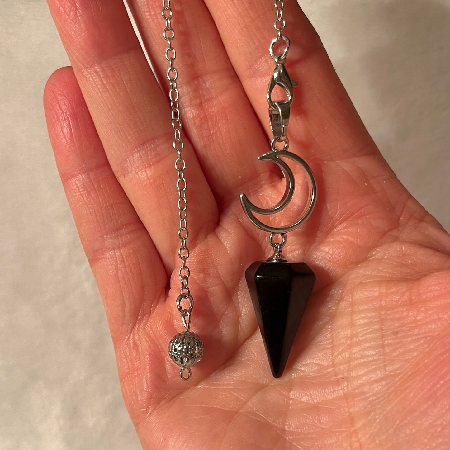 1 inch black obsidian pendulum attarched to a silver crescent moon, both connected to an 8 inch silver chain, displayed delicately in the palm of a hand