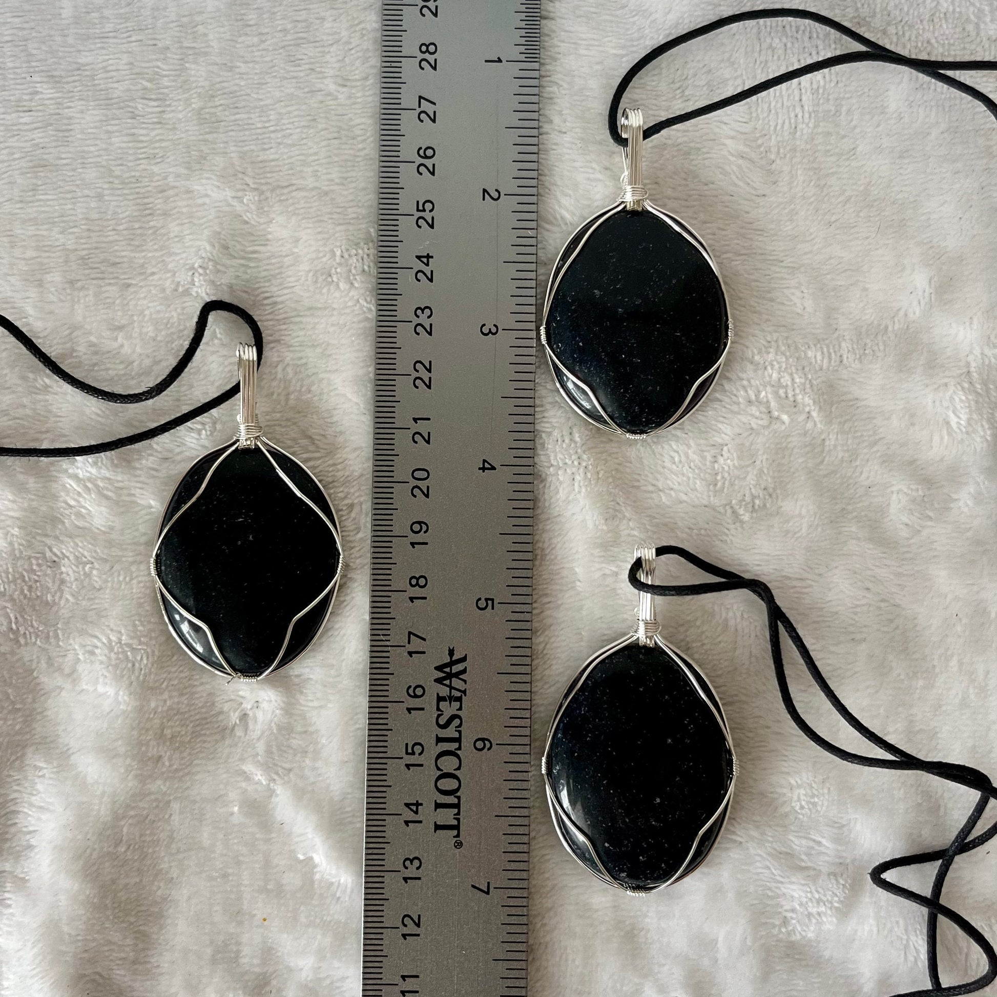 3 obsidian worry stones beautifully framed by silver wire and made into pendants, with adjustable black cords, displayed next to a ruler.  obsidian crystal worry stones are approximately 1 1/2" long