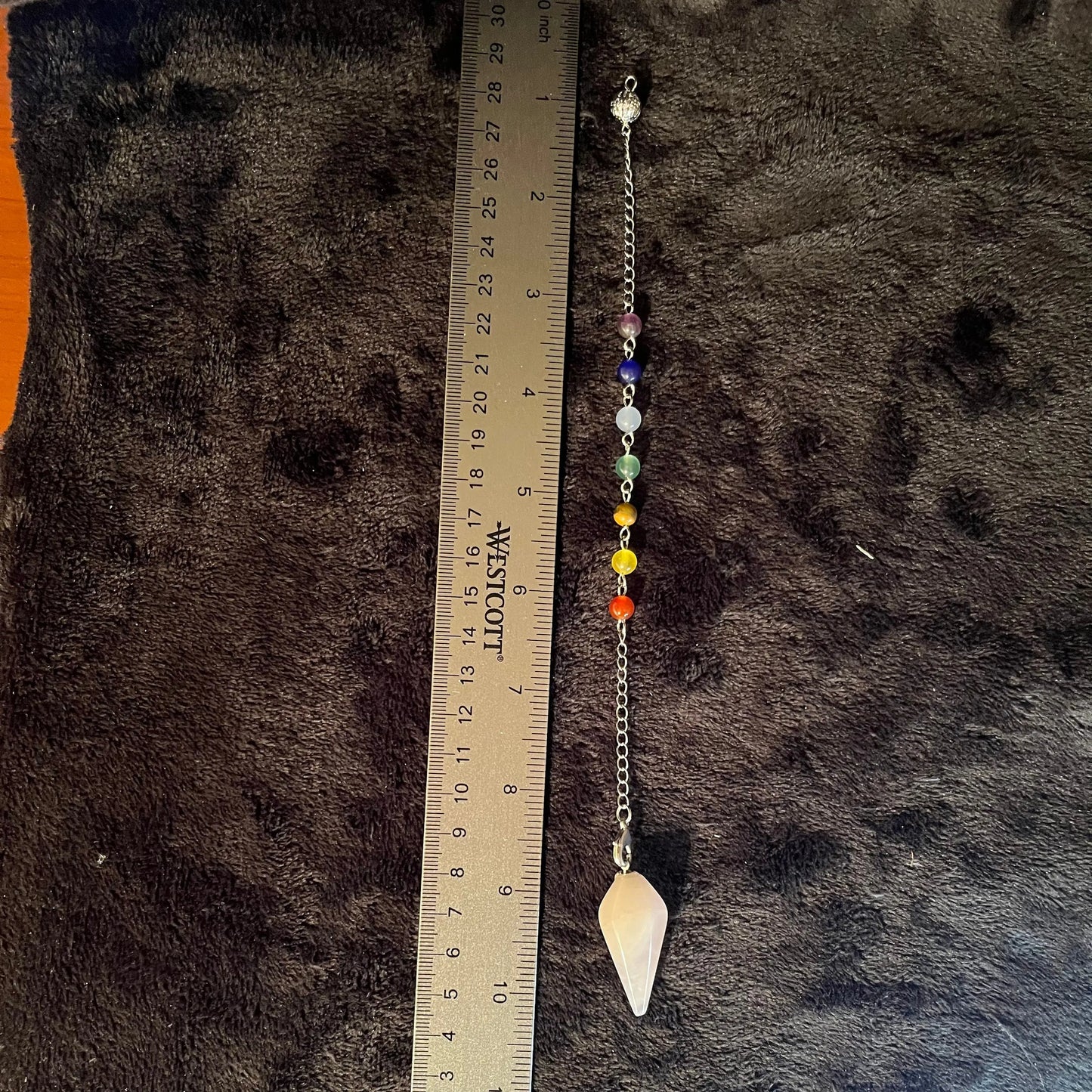 Close up of rose quartz pendulum attatched to a chain containing chakra beads, next to a ruler 